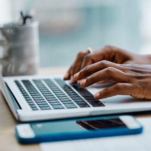 Person tippt auf einem Laptop. Neben ihr liegt ein Smartphone.