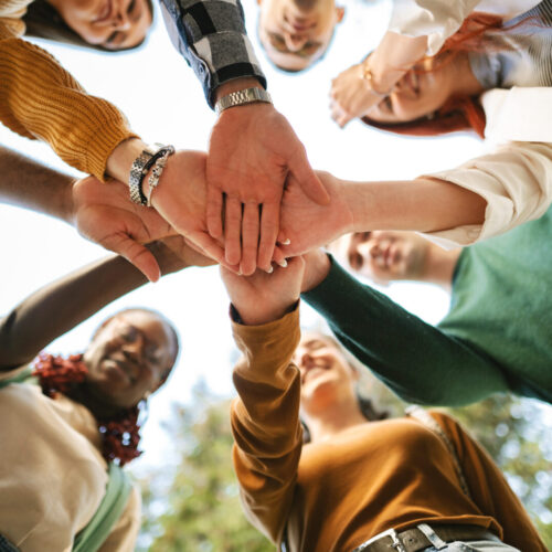Eine Gruppe von Personen hält in der Mitte des Bildes ihre Händer übereinander.